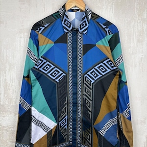 May include: A long-sleeved shirt with a geometric pattern in shades of blue, teal, brown, and black. The shirt is hanging on a wooden hanger. The design includes squares, triangles, and rectangular shapes. The shirt has a collared neckline.