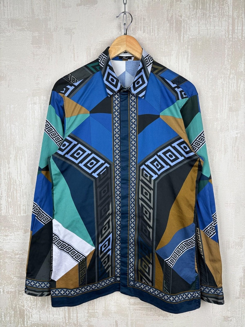 May include: A long-sleeved shirt with a geometric pattern in shades of blue, teal, brown, and black. The shirt is hanging on a wooden hanger. The design includes squares, triangles, and rectangular shapes. The shirt has a collared neckline.