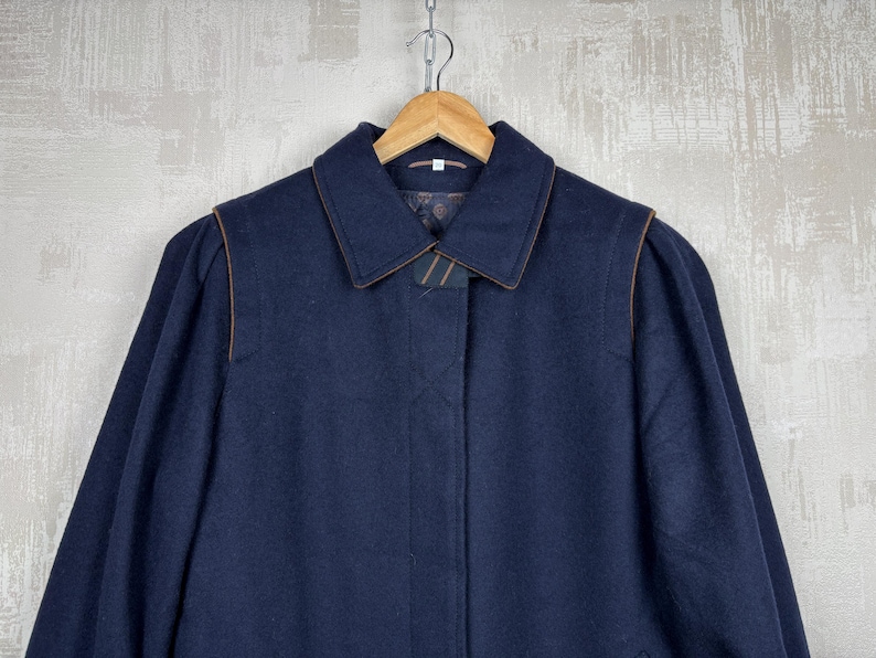 May include: A navy blue wool coat with a brown trim around the collar and pockets. The coat has a long sleeve and a button closure.