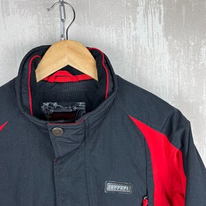 May include: Black jacket with red accents, hanging on a wooden hanger. The jacket features a high collar with red trim, a button closure, and a small rectangular patch with text. Red panels are on the shoulders and sides.