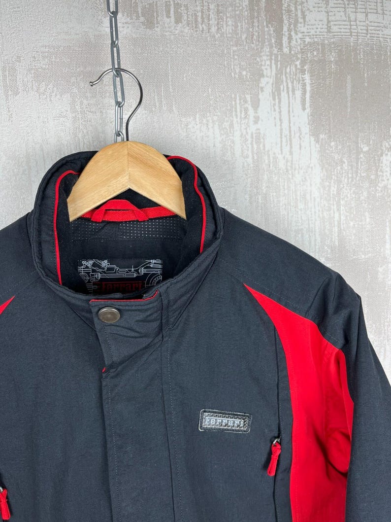 May include: Black jacket with red accents, hanging on a wooden hanger. The jacket features a high collar with red trim, a button closure, and a small rectangular patch with text. Red panels are on the shoulders and sides.