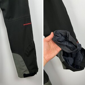 May include: Black ski pants with a gray panel on the lower leg. The pants have a zipper on the lower leg and the word "MAMMUT" is embroidered on the right leg.