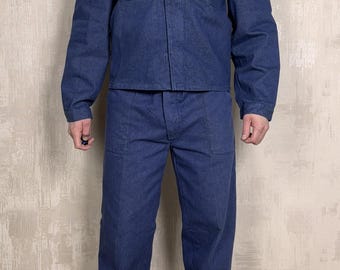 May include: A denim workwear set in a medium blue hue. The set includes a collared jacket with two chest pockets and matching pants. The model is wearing gray sneakers. The outfit is a classic workwear style.