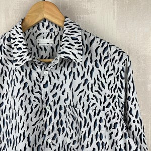 May include: A white long-sleeved button-down shirt with a black and blue abstract animal print. The shirt has a single chest pocket.