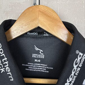 May include: Black collared shirt with white embroidered text and logos. The shirt features the Newcastle Falcons logo and the text "KooGa MADE FOR RUGBY". The shirt is hanging on a wooden hanger.