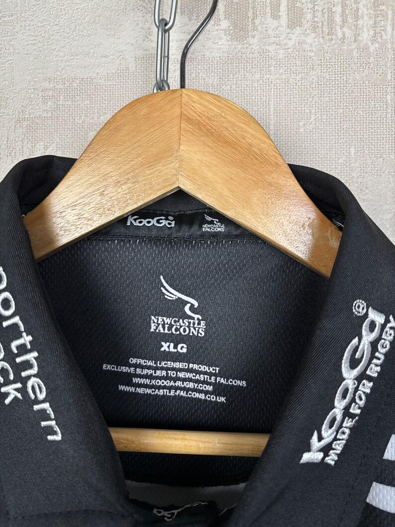 May include: Black collared shirt with white embroidered text and logos. The shirt features the Newcastle Falcons logo and the text "KooGa MADE FOR RUGBY". The shirt is hanging on a wooden hanger.