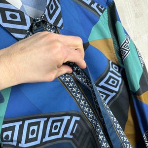 May include: A patterned button-down shirt in shades of blue, teal, black, and brown. The shirt has a collar and long sleeves. The design incorporates diamond and square shapes. The shirt is made of cotton.