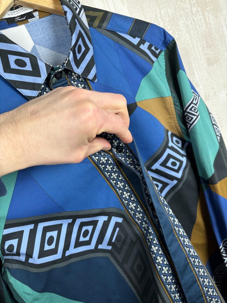 May include: A patterned button-down shirt in shades of blue, teal, black, and brown. The shirt has a collar and long sleeves. The design incorporates diamond and square shapes. The shirt is made of cotton.