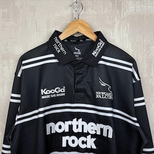 May include: Black and white rugby jersey with the words "northern rock" in large white letters. The jersey has white stripes and the "Newcastle Falcons" logo. The collar has the words "northern rock" in white.