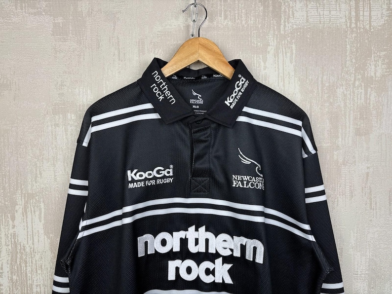 May include: Black and white rugby jersey with the words "northern rock" in large white letters. The jersey has white stripes and the "Newcastle Falcons" logo. The collar has the words "northern rock" in white.