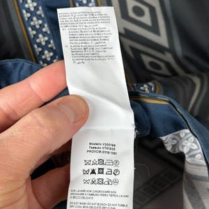 May include: A garment label showing care symbols and fabric details. The label is white with black text and is attached to a patterned fabric with blue, grey, and white geometric designs.