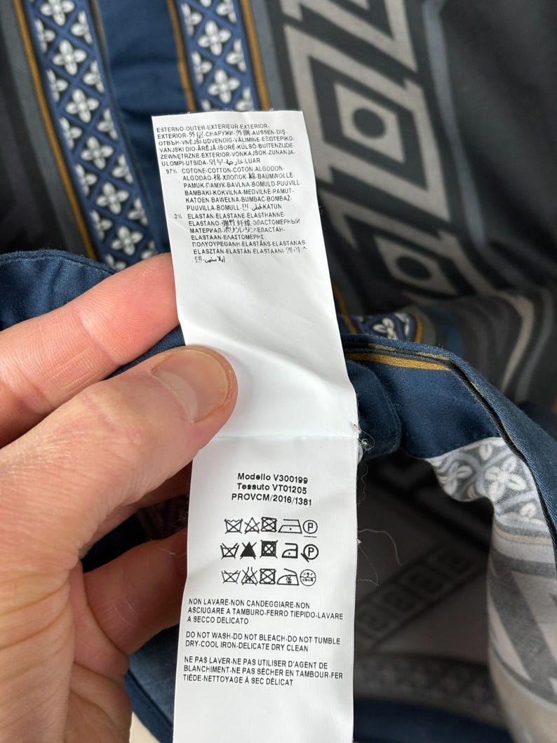 May include: A garment label showing care symbols and fabric details. The label is white with black text and is attached to a patterned fabric with blue, grey, and white geometric designs.
