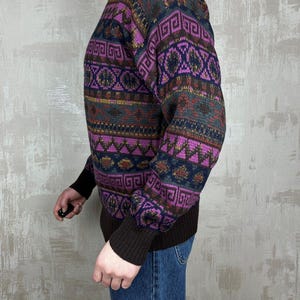 K&ouml;nnte beinhalten: Ein gemusterter Pullover in Lila-, Braun- und Marineblaut&ouml;nen. Der Pullover hat ein geometrisches Design auf Brust und &Auml;rmeln. Die Manschetten und der Kragen sind einfarbig braun. Die Person tr&auml;gt eine blaue Jeans.