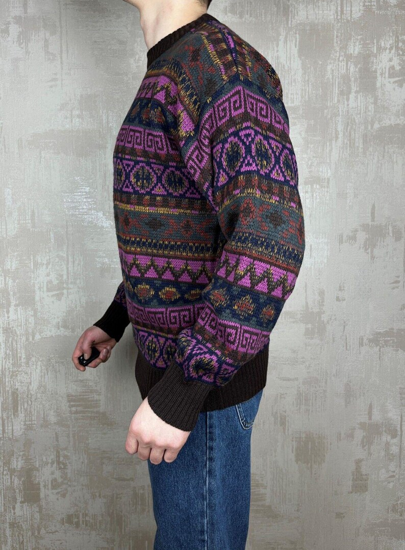K&ouml;nnte beinhalten: Ein gemusterter Pullover in Lila-, Braun- und Marineblaut&ouml;nen. Der Pullover hat ein geometrisches Design auf Brust und &Auml;rmeln. Die Manschetten und der Kragen sind einfarbig braun. Die Person tr&auml;gt eine blaue Jeans.