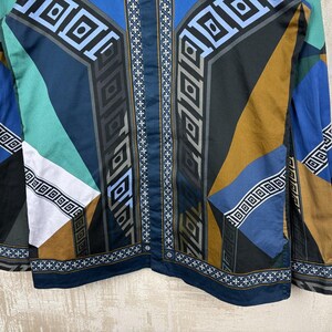 May include: A long-sleeved shirt with a geometric pattern in shades of blue, teal, brown, and black. The design features repeating square and rectangular shapes, with a central vertical stripe of white crosses.