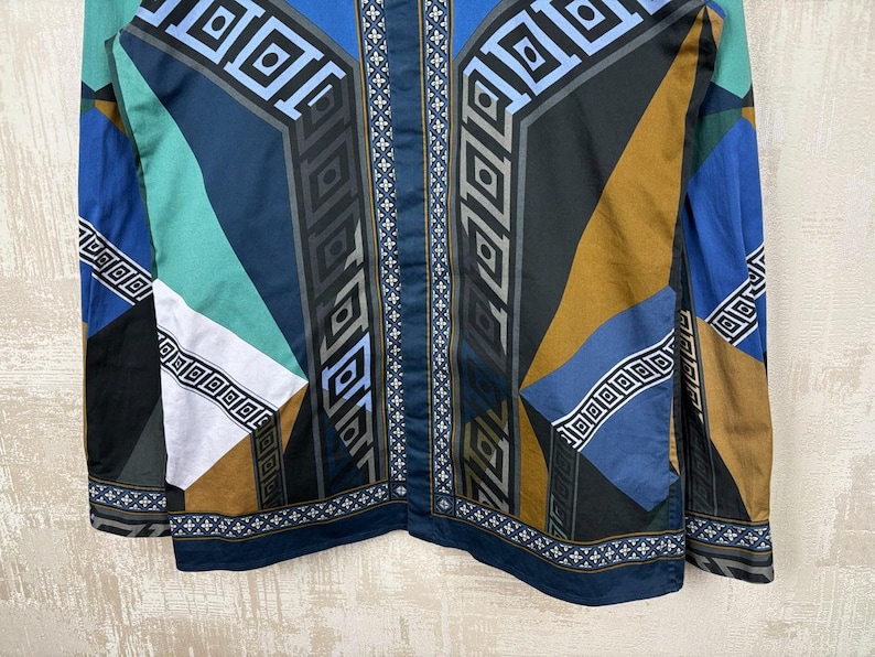 May include: A long-sleeved shirt with a geometric pattern in shades of blue, teal, brown, and black. The design features repeating square and rectangular shapes, with a central vertical stripe of white crosses.