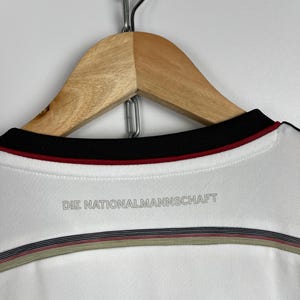May include: White soccer jersey with black and red trim around the neck. The back of the jersey has the text "DIE NATIONALMANNSCHAFT" printed in gray.