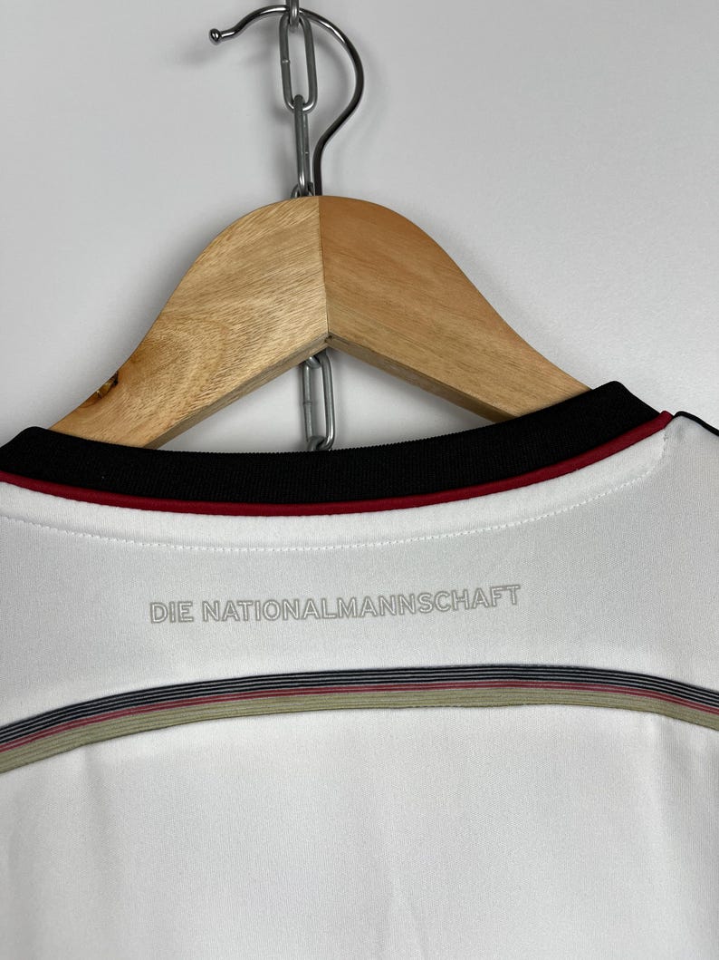 May include: White soccer jersey with black and red trim around the neck. The back of the jersey has the text "DIE NATIONALMANNSCHAFT" printed in gray.