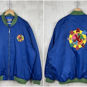 May include: A blue bomber jacket with green trim. The front has a small embroidered patch, while the back features a large circular design with the text "NMC NATURAL 1973". The jacket is hanging on a wooden hanger.