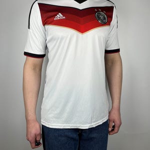 May include: White soccer jersey with black and red stripes, the Adidas logo, and the German national team crest. The jersey has a V-neck and short sleeves.