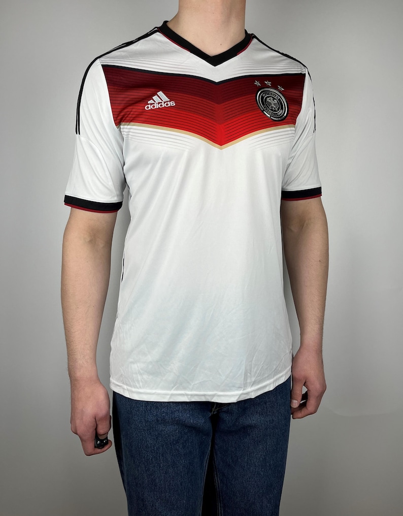 May include: White soccer jersey with black and red stripes, the Adidas logo, and the German national team crest. The jersey has a V-neck and short sleeves.