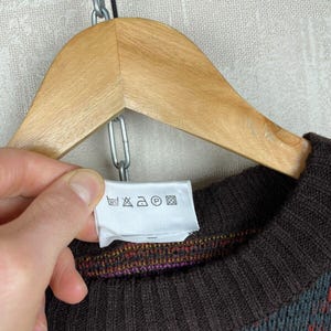 K&ouml;nnte beinhalten: Ein braun-blau gemusterter Pullover mit geripptem Kragen und B&uuml;ndchen, der an einem Holzb&uuml;gel h&auml;ngt. Der Pullover hat ein farbenfrohes Design und ein Etikett mit Pflegehinweisen.