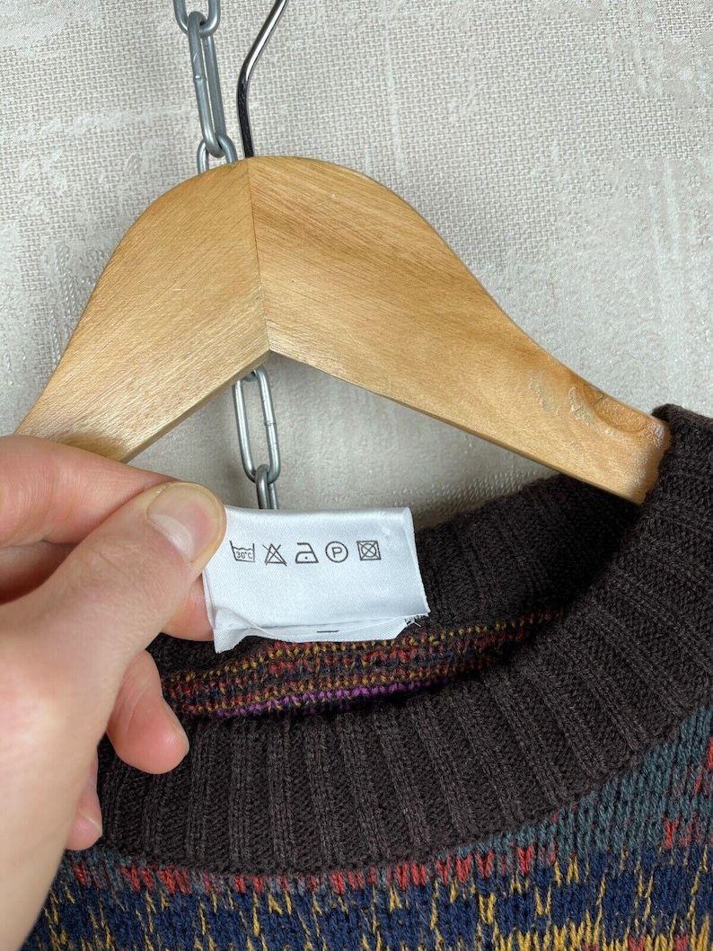 K&ouml;nnte beinhalten: Ein braun-blau gemusterter Pullover mit geripptem Kragen und B&uuml;ndchen, der an einem Holzb&uuml;gel h&auml;ngt. Der Pullover hat ein farbenfrohes Design und ein Etikett mit Pflegehinweisen.