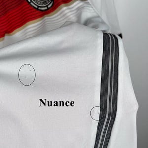 May include: White soccer jersey with black and white stripes on the sleeve. The jersey has the German national team logo on the chest and the word "Nuance" written on the sleeve.