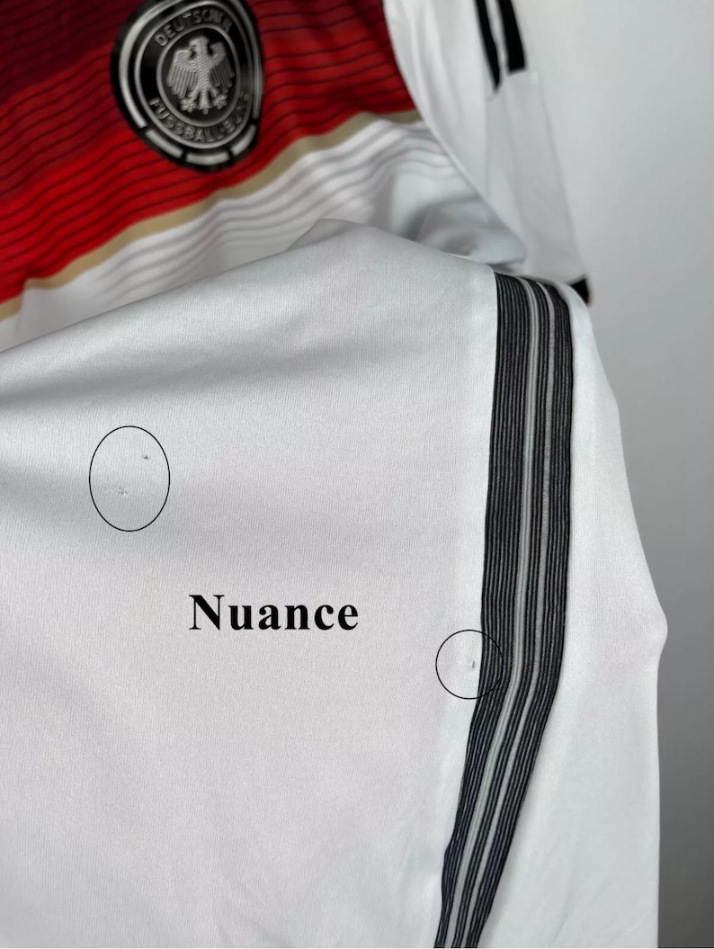 May include: White soccer jersey with black and white stripes on the sleeve. The jersey has the German national team logo on the chest and the word "Nuance" written on the sleeve.