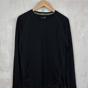 May include: Black long-sleeve shirt hanging on a wooden hanger. The shirt has a crew neck with a thin, multi-colored stripe detail. The seams are visible with a lighter thread. The brand logo is visible on the upper chest.