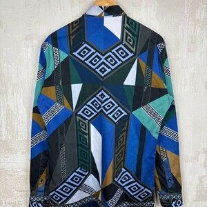 May include: A long-sleeved shirt with a geometric pattern in blue, green, brown, and white. The shirt is hanging on a wooden hanger. The design features squares and diamond shapes. The shirt's design is a striking, bold statement.