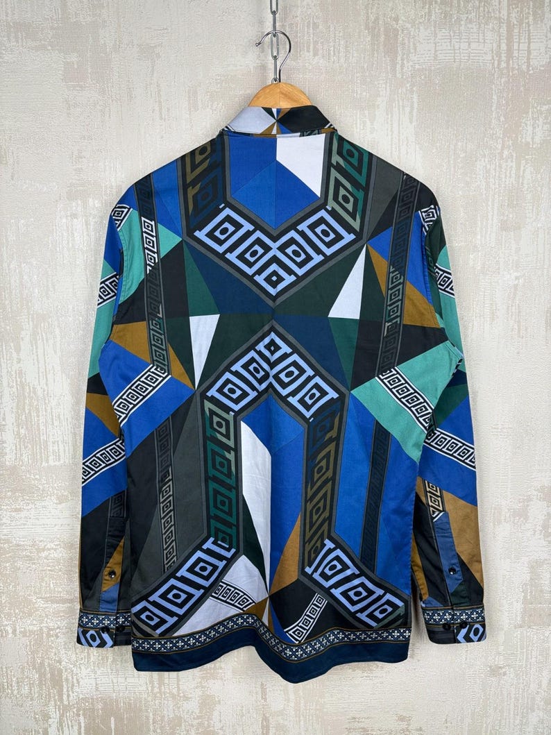May include: A long-sleeved shirt with a geometric pattern in blue, green, brown, and white. The shirt is hanging on a wooden hanger. The design features squares and diamond shapes. The shirt's design is a striking, bold statement.