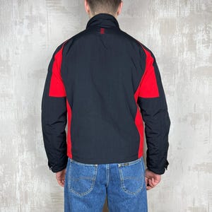 May include: Black and red jacket with a high collar. The jacket has red panels on the shoulders and sides, with a black body and sleeves. The jacket is worn with blue jeans.