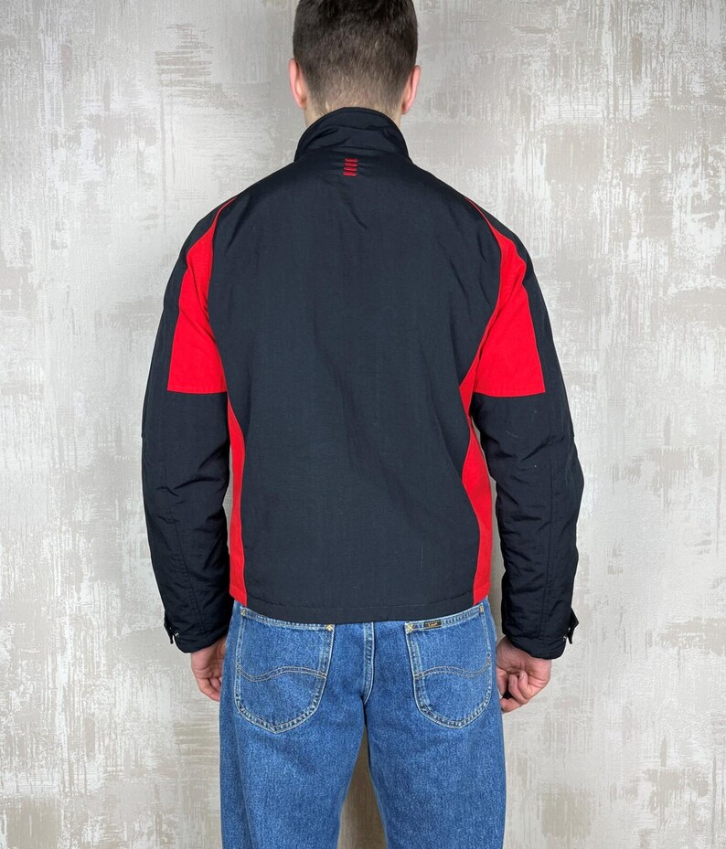 May include: Black and red jacket with a high collar. The jacket has red panels on the shoulders and sides, with a black body and sleeves. The jacket is worn with blue jeans.