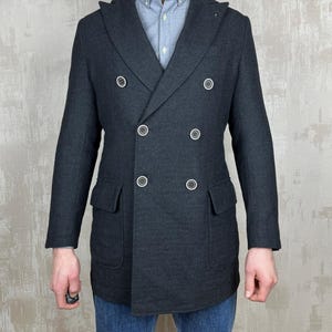 May include: A dark gray double-breasted coat with six white buttons and two front pockets. The coat is worn over a light blue button-down shirt and blue jeans. The coat has a classic, tailored design.