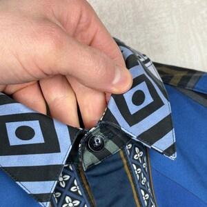 May include: Close-up of a blue button-down shirt. The collar has a geometric pattern, and the placket features a floral design. The shirt has a black button and a blue and black geometric design.