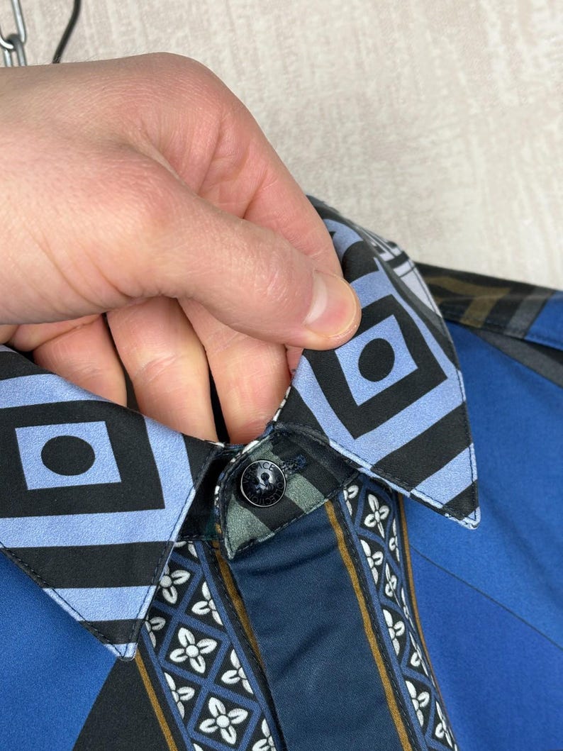 May include: Close-up of a blue button-down shirt. The collar has a geometric pattern, and the placket features a floral design. The shirt has a black button and a blue and black geometric design.
