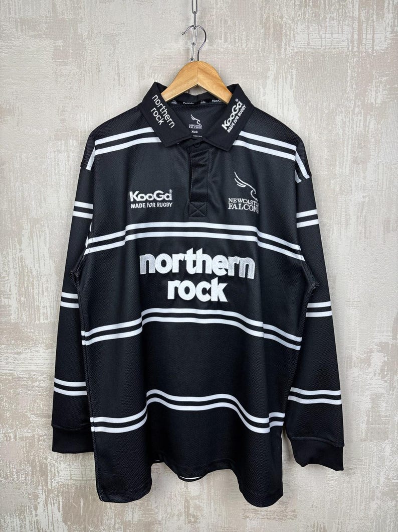 May include: Black long-sleeve rugby shirt with white horizontal stripes and the text "northern rock" in large white letters. The shirt also features the "Newcastle Falcons" logo and "KooGa MADE FOR RUGBY" text.