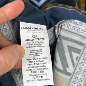 May include: A close-up of a Gianni Versace garment label. The label displays the brand name, address, and product details. The fabric features a geometric design in blue, grey, and beige tones.