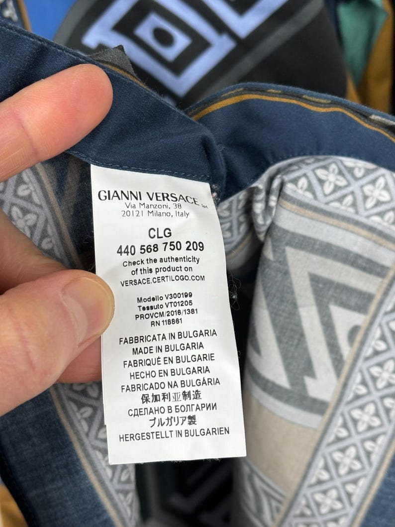 May include: A close-up of a Gianni Versace garment label. The label displays the brand name, address, and product details. The fabric features a geometric design in blue, grey, and beige tones.
