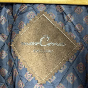 May include: A brown suede patch with the text "marCora" and "EXCLUSIV" in white lettering. The patch is diamond-shaped with scalloped edges.