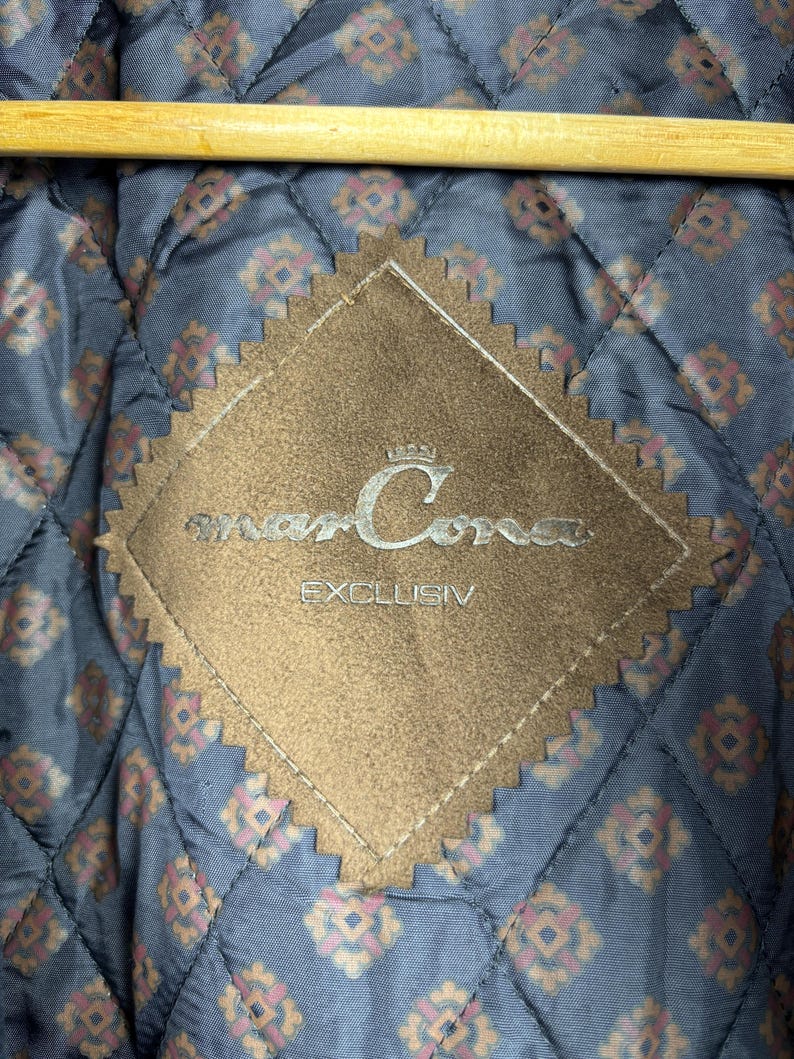 May include: A brown suede patch with the text "marCora" and "EXCLUSIV" in white lettering. The patch is diamond-shaped with scalloped edges.