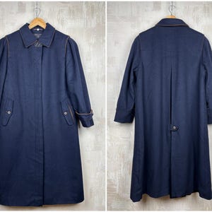 May include: A navy blue wool coat with a double breasted closure, two pockets, and a brown leather trim. The coat has a long, straight silhouette and a collar.