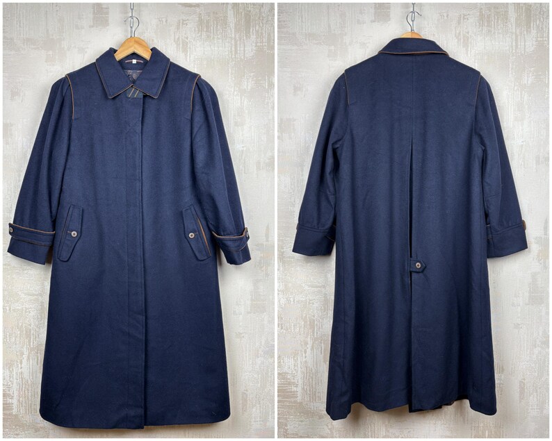 May include: A navy blue wool coat with a double breasted closure, two pockets, and a brown leather trim. The coat has a long, straight silhouette and a collar.