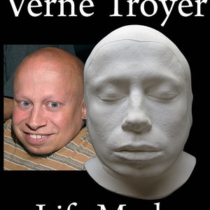 Puede incluir: Una máscara mortuoria de yeso blanco de Verne Troyer, un actor estadounidense. La máscara muestra un primer plano de su rostro con los ojos cerrados. La máscara está sobre una superficie negra. El texto "Verne Troyer" y "Life Mask" está impreso debajo de la máscara.