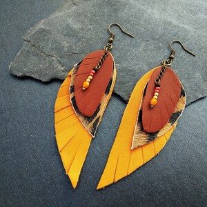 May include: A pair of dangle earrings with a layered feather design. The feathers are made of leather in shades of yellow, brown, and black. The earrings are accented with small orange and red beads.