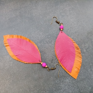 May include: A pair of pink and orange leather feather earrings. The earrings are made of two layers of leather, with the top layer being pink and the bottom layer being orange. The earrings are fringed at the bottom and have a small pink bead at the top.
