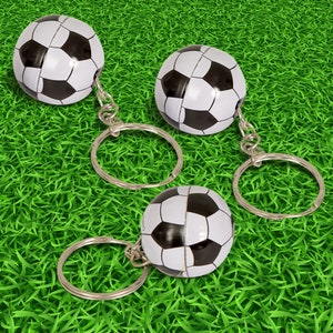 May include: Three black and white soccer ball keychains with silver key rings. The keychains are on a green grass background.