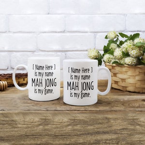 May include: Two white ceramic mugs with black text. The text on the mugs reads "[Name Here] is my name MAH JONG is my game."