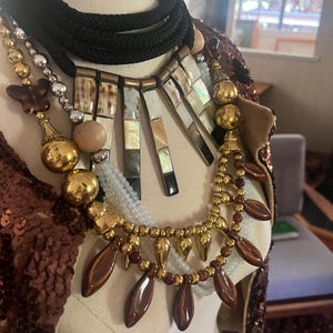 Könnte beinhalten: Eine mehrreihige Halskette mit einem schwarzen Seil-Choker, goldenen Perlen und braunen und weißen rechteckigen Anhängern. Die Halskette hat auch braune tropfenförmige Perlen und einen goldfarbenen Anhänger.